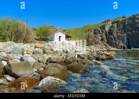 Traditionelle weiße Kirche auf einem Kiesstrand mit transparentem türkisfarbenem Wasser und Küstenhügeln in Skala Eressos auf der Insel Lesbos, Griechenland Stockfoto
