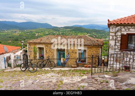 Molyvos, Griechenland - 22. April 2026: Traditionelle Steinhäuser mit Ziegeldächern und blauen Akzenten säumen eine malerische Kopfsteinpflasterstraße in Molyvos, Griechenland Stockfoto