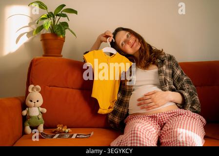 Werdende Mutter sitzt auf dem Sofa, hält den Babybody, feiert neues Leben und zukünftige Ankunft, symbolisiert mütterliche Liebe, Schwangerschaftsreise und Familienplanung zu Hause Stockfoto
