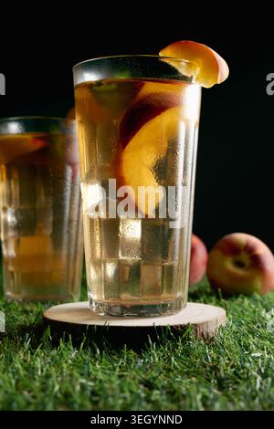 Nahaufnahme von zwei erfrischenden Gläsern Pfirsicheistee mit Fruchtscheiben, die auf hölzernen Untersetzern über grünem Gras und dunklem Hintergrund stehen Stockfoto