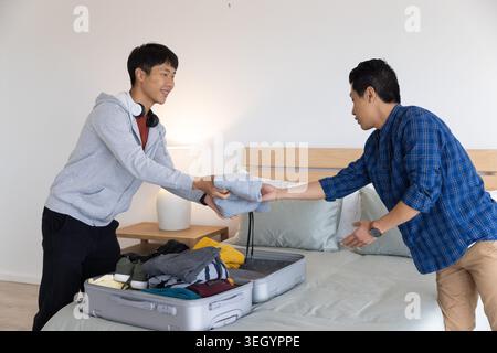 Asiatische Männer packen offenen Koffer auf dem Bett, ziehen gefalteten Pullover, während sie Kopfhörer und Uhr tragen Stockfoto