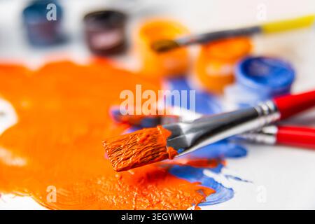 Hellorange und blaue Farbe bedeckt eine Palette mit Pinseln in der Nähe. Künstler kreieren an einem Tisch, der mit Farbzubehör gefüllt ist. Stockfoto