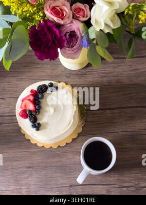 Eine Person sitzt an einem Holztisch mit einem Kuchen mit Beeren und einer Tasse Kaffee. In der Nähe befindet sich eine Vase mit bunten Blumen, die zur Kulisse beiträgt. Stockfoto