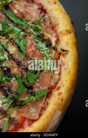 Eine frische Pizza liegt auf einer schwarzen Oberfläche, gekrönt mit Schinkenscheiben, Rucola und Balsamico-Glasur. Die Kruste ist goldfarben und knusprig Stockfoto