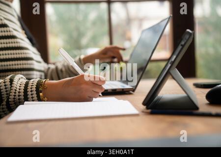 Moderner, professioneller Arbeitsbereich, in dem eine Frau beim Schreiben von Notizen mit Laptop und Tablet spielt. Ideal für Unternehmen, hybride Arbeit, Technologie und Produktivität Stockfoto