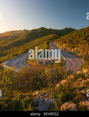 Aus der Vogelperspektive auf eine gewundene Bergstraße mit einer scharfen Haarnadelkurve und einem Steindenkmal umgeben von üppigen grünen Hügeln während der goldenen Stunde in Rijeka, Pri Stockfoto