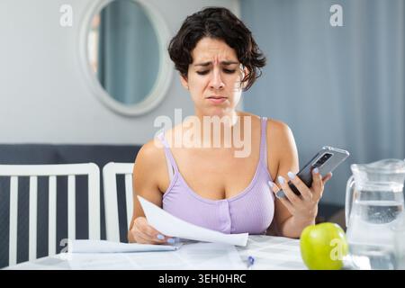 Schockierte junge Frau, die Dokumente ansieht und Smartphone in der Hand hält Stockfoto
