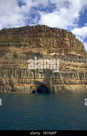 Ein vertikaler Niedrigwinkelschuss aus dem Wasser, der geschichtete vulkanische Gesteinsschichten auf einer massiven Küstenklippe mit einer natürlichen Meereshöhle am Fuß zeigt. Stockfoto