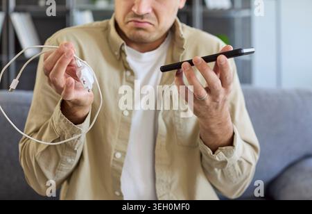 Nahaufnahme eines Mannes mit Telefon und Ladegerät, verwirrt durch Ladefehler zu Hause Stockfoto