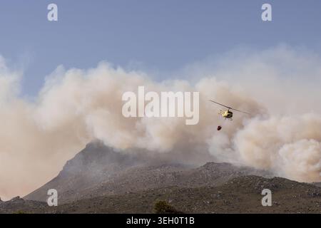 Blick auf einen gelben Feuerlöschhubschrauber mit einem Wassereimer in der Nähe von dickem Rauch, der von einem Waldbrand an den Hängen des Tafelbergs Kapstadt, We, aufsteigt Stockfoto