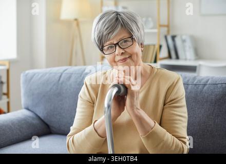 Porträt einer lächelnden älteren Frau, die zu Hause sitzt und einen Gehstock hält und in die Kamera schaut Stockfoto