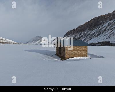 KAZA, Indien - 28. Januar 2026: Blick auf eine kleine Steinhütte mit gewelltem Metalldach in einer riesigen schneebedeckten Landschaft des Spiti Valley, Surro Stockfoto