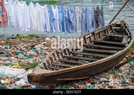 Dhaka Division, Bangladesch - 14. Mai 2026: Blick auf ein hölzernes Boot auf den mit Müll gefüllten Ufern des Buriganga Flusses mit Wäsche im Hinterland Stockfoto