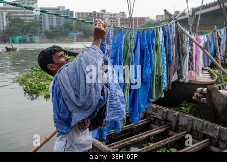 Dhaka Division, Bangladesch - 14. Mai 2026: Blick auf einen Mann, der seine Wäsche an einem Seil über einem Holzboot auf dem Buriganga River mit städtischen Gebäuden in Th hängt Stockfoto