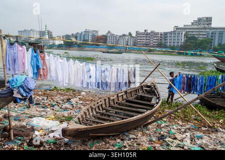 Dhaka Division, Bangladesch - 14. Mai 2026: Blick auf den Buriganga River mit einem Mann, der an den Schlangen neben einem Holzboot auf einem verschmutzten Ufer seine Wäsche hängt. Stockfoto