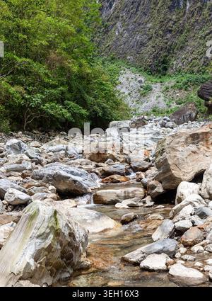 Sanzhanxi felsiges Flussbett im Taroko-Nationalpark. Große Felsbrocken säumen flaches Wasser. Grüner Wald bedeckt steile Berghänge. Stockfoto