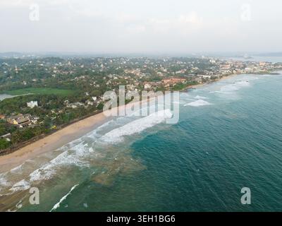 Blick aus der Vogelperspektive auf die Küste des Indischen Ozeans mit Wellen, die am Sandstrand zusammenbrechen, und städtische Gebäude, umgeben von üppigem Grün in Galle, Southern PR Stockfoto
