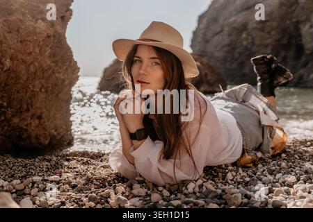Frau Strand Kieselsteine junge Frau mit Strohhut liegt an der Küste mit felsigen Klippen während der goldenen Stunde Stockfoto