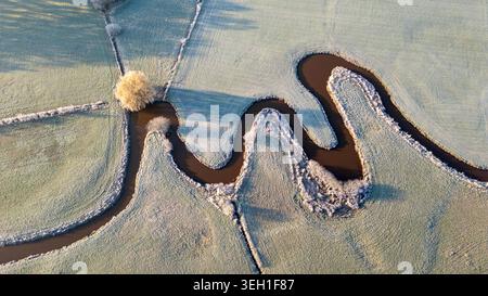 Aus der Vogelperspektive eines mäandernden Baches, der durch frostbedeckte Wiesen mit einem einsamen goldenen Baum in de Schiphorst, Drenthe, Niederlande fließt. Stockfoto