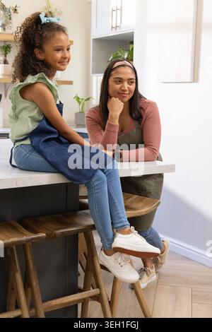 Vielfältige weibliche Mutter und afroamerikanische Jugendliche sitzen auf der Kücheninsel in Olivgrün-Schürzen Stockfoto