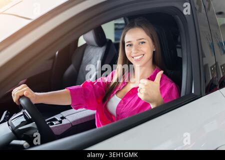 Glückliche junge Frau, die im Auto sitzt und im Autohändler die Daumen hochlegt Stockfoto