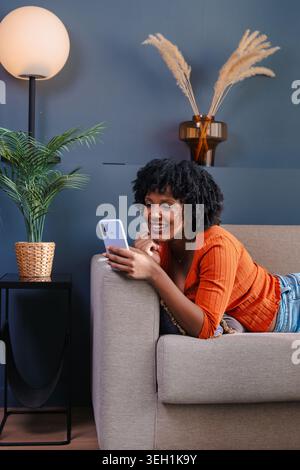 Vertikale Aufnahme, die auf dem Sofa liegt und zu Hause ihr Smartphone benutzt Stockfoto