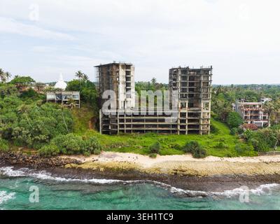 Aus der Vogelperspektive eines unfertigen Betonbauwerks an der Küste mit einer weißen Stupa und tropischem Grün in Mirissa, Südprovinz, Sri Stockfoto