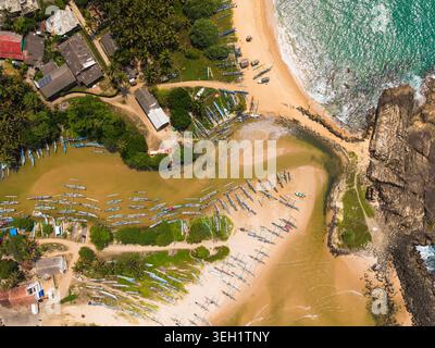 Blick aus der Vogelperspektive auf die traditionellen Auslegerboote, die am Sandstrand und an der Lagune von Dodanduwa, umgeben von Palmen und Küstengebäuden, angereiht sind; Stockfoto