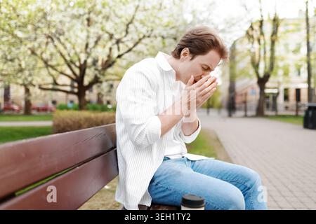 Ein Mann, der an Frühlingsallergien leidet, niest in die Hände, während er auf einer Parkbank sitzt, umgeben von blühenden Bäumen. Saisonales Heuschnupfen-Konzept. H Stockfoto