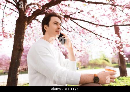 Lächelnder Freelancer, der auf einem Smartphone spricht und Kaffee hält, während er draußen unter blühenden rosa Frühlingskirschblüten sitzt. Remote Work Life Stockfoto