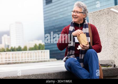 Reifer Mann sitzt auf Betonvorsprung in rotem Pullover und Schal, der die Take-Out-Box hält, Kopierraum Stockfoto