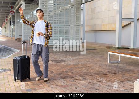 Asiatischer Mann, der auf der Transitplattform steht, mit schwarzem Koffer und Kaffee, mit gelbem Karo Stockfoto