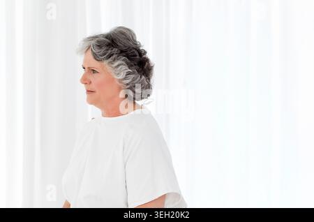 Eine ältere Frau, die im Profil steht und ein weißes Kleid trägt, sieht von transparenten Vorhängen links, Kopierraum Stockfoto