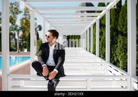 Hübscher Mann in Anzug und Sonnenbrille, der auf der Treppe sitzt und wegblickt Stockfoto
