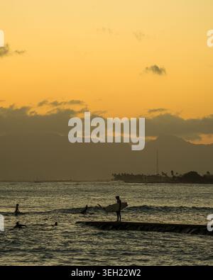 Honolulu, Hawaii, USA - 19. Mai 2025: Ein surfer in Silhouette steht bei Sonnenuntergang auf einem Steg, während mehrere Surfer im Wasser vor Waikiki warten. Stockfoto
