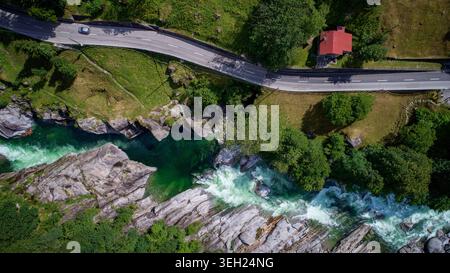 Aus der Vogelperspektive auf den türkisfarbenen Fluss Verzasca, der durch felsige Ufer fließt, neben einer gewundenen Bergstraße und einem Haus mit rotem Dach im Valle Verzasca, Stockfoto