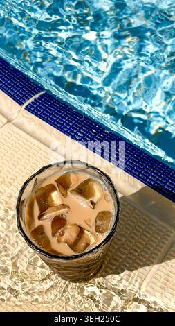 Eiskaffee mit Eiswürfeln in einem Glas neben einem Swimmingpool mit klarem blauem Wasser, das an einem warmen Tag das Sonnenlicht reflektiert Stockfoto