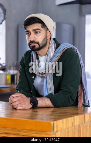 Mann lehnt sich auf Holztheke in einem modernen Café, trägt Mütze und langen mehrfarbigen Schal. Bart, Hut, Uhr, Armband, Porträt, solo, kontemplativ Stockfoto