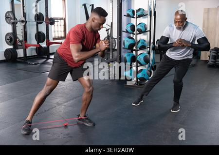 Männlicher Trainer und Kunde, der seitliche Schritte mit rotem Schleifenband im Fitnessstudio mit rotem Hemd durchführt. Fitness, Coach, Athleten, Bewegung, Mobilität, Stärke, Gewichte Stockfoto