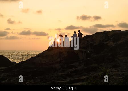 Blick auf Silhouetten von Menschen, die auf felsigen Klippen am Meer sitzen, während eines goldenen Sonnenuntergangs in Galle, Südprovinz, Sri Lanka. Stockfoto