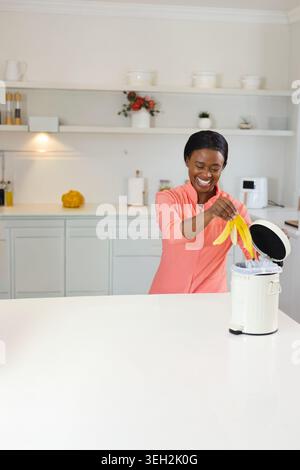 Afroamerikanerin in Korallenoberteil, die Bananenschalen über dem Treteimer hält, an der Haustheke, im Kopierraum Stockfoto