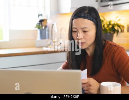 Chinesin in Rostspitze, Kopfband mit Polka-Dot, die am Laptop arbeitet und Papier an der Küchentheke liest Stockfoto