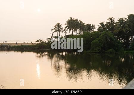 Blick auf eine ruhige Lagune, die den goldenen Sonnenuntergang mit üppigen Palmen und Menschen am fernen Strand in Galle, Südprovinz, Sri Lanka reflektiert. Stockfoto