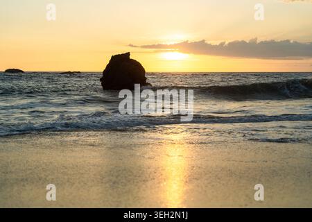 Blick auf eine große Felsformation im Ozean während eines goldenen Sonnenuntergangs mit Wellen, die auf den Sandstrand in Galle, Südprovinz, Sri Lanka stürzen. Stockfoto