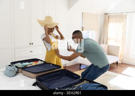 Afroamerikanisches Paar packt im Schlafzimmer, eine Frau versucht einen Sonnenhut, ein Mann, der ein Foto vom Telefon macht, Koffer Stockfoto