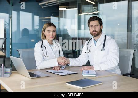 Zwei verschiedene medizinische Fachkräfte in Laborkitteln und Stethoskopen schütteln in einem modernen Büro die Hand über einen Schreibtisch und symbolisieren Partnerschaft, Vertrauen und kollaborative Entscheidungen im Gesundheitswesen Stockfoto