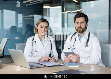 Zwei medizinische Fachkräfte, ein Mann und eine Frau in Labormänteln und Stethoskopen, die Dokumente und digitale Informationen sorgfältig überprüfen, während sie an einem Schreibtisch in einem modernen Krankenhausbüro sitzen Stockfoto