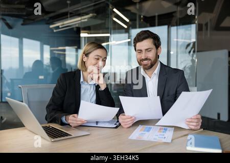 Zwei lächelnde Geschäftskollegen prüfen Finanzberichte und Papiere, verwenden einen Laptop und arbeiten an einem Projekt in einer modernen Büroumgebung zusammen Stockfoto