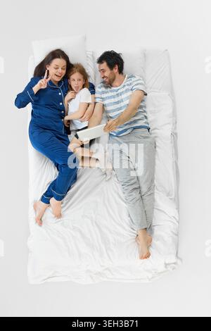 Verspielte Familie, die mit fröhlicher Tochter und entspannten Eltern ein Tablet-Selfie auf dem Bett macht. Stockfoto