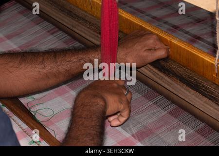 Nahaufnahme eines indischen Handwebhandwerkers, der mit rosa und weiß karierten Jamdani-Textilien nach traditionellen Webtechniken auf einem Holzwebstuhl arbeitet. Stockfoto
